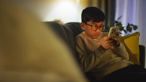 Young-Boy-Sitting-On-Sofa-In-Lounge-At-Home-Playing-Game-On-Mobile-Phone-At-Night-3